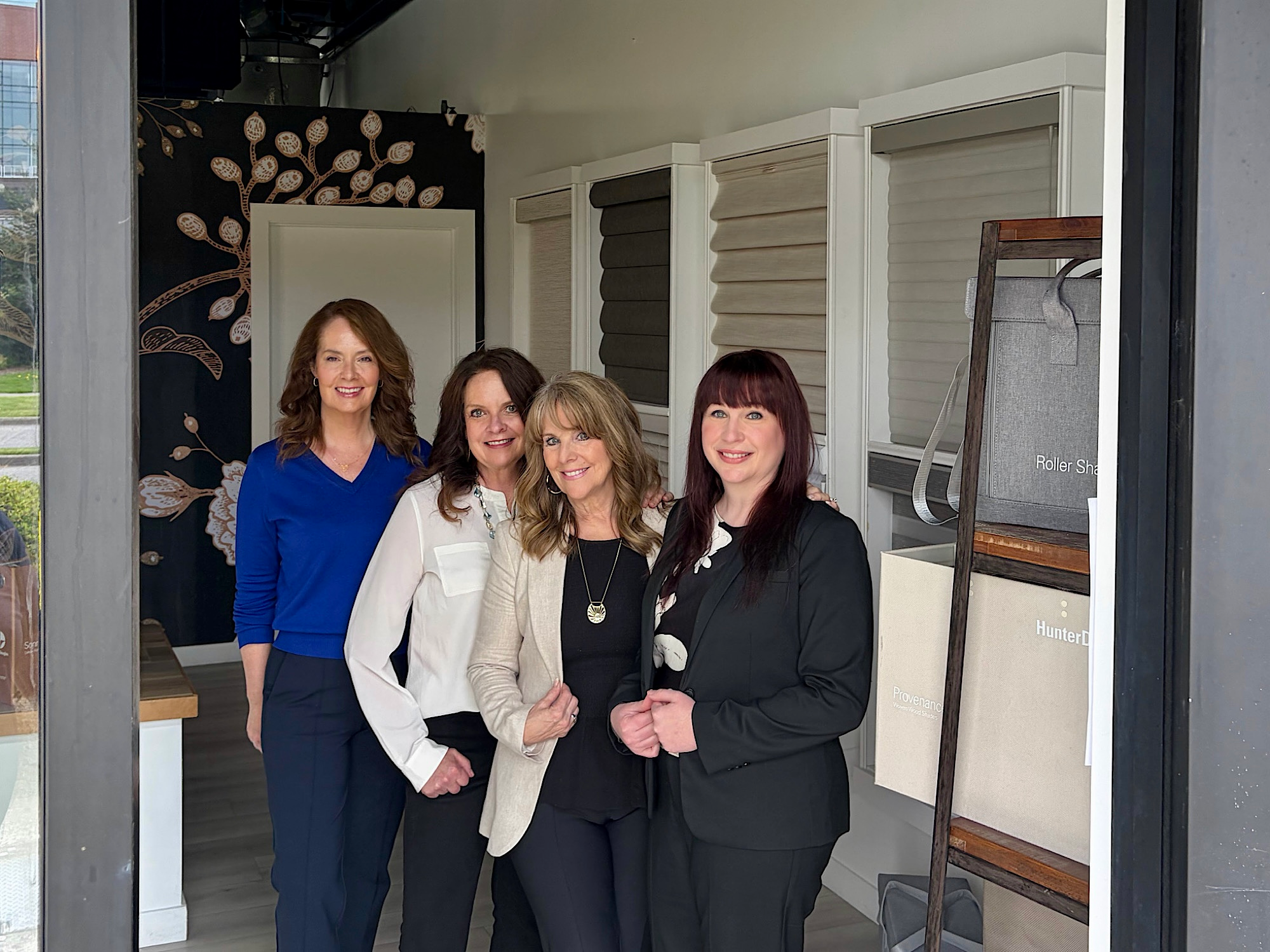 Four smiling women posing together in a window treatment showroom with roller shades and blind samples on display behind them.