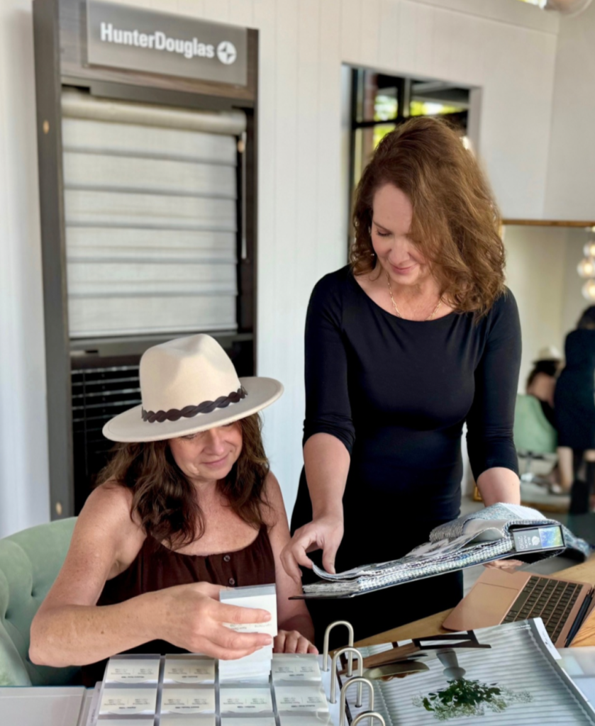 Woman in a beige hat sits at a desk, while a second woman in black guides her through fabric swatches, with a Hunter Douglas display in the background.