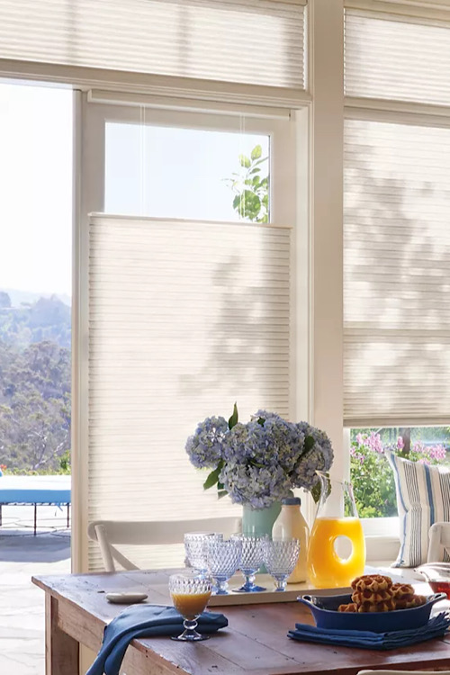 Sunlit dining table by large windows with white blinds; blue hydrangea centerpiece, glassware, orange juice jug, and waffles on a blue plate.