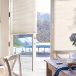 Honeycomb shades raised on glass doors and windows in a sunny dining area overlooking a garden