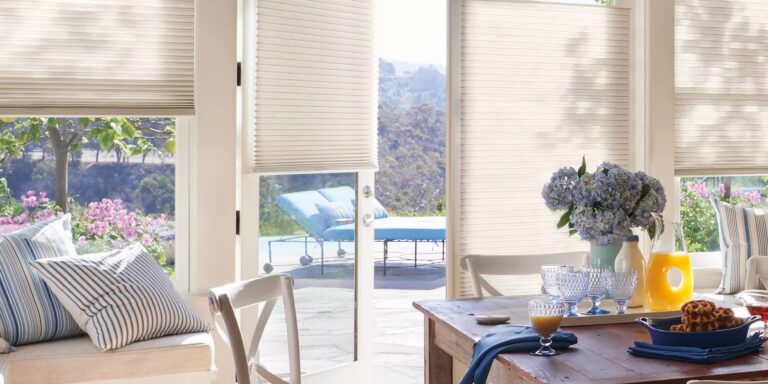 Honeycomb shades raised on glass doors and windows in a sunny dining area overlooking a garden