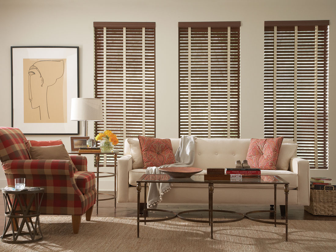 Three dark brown faux wood blinds with light cloth tapes above a white sofa and a red plaid armchair