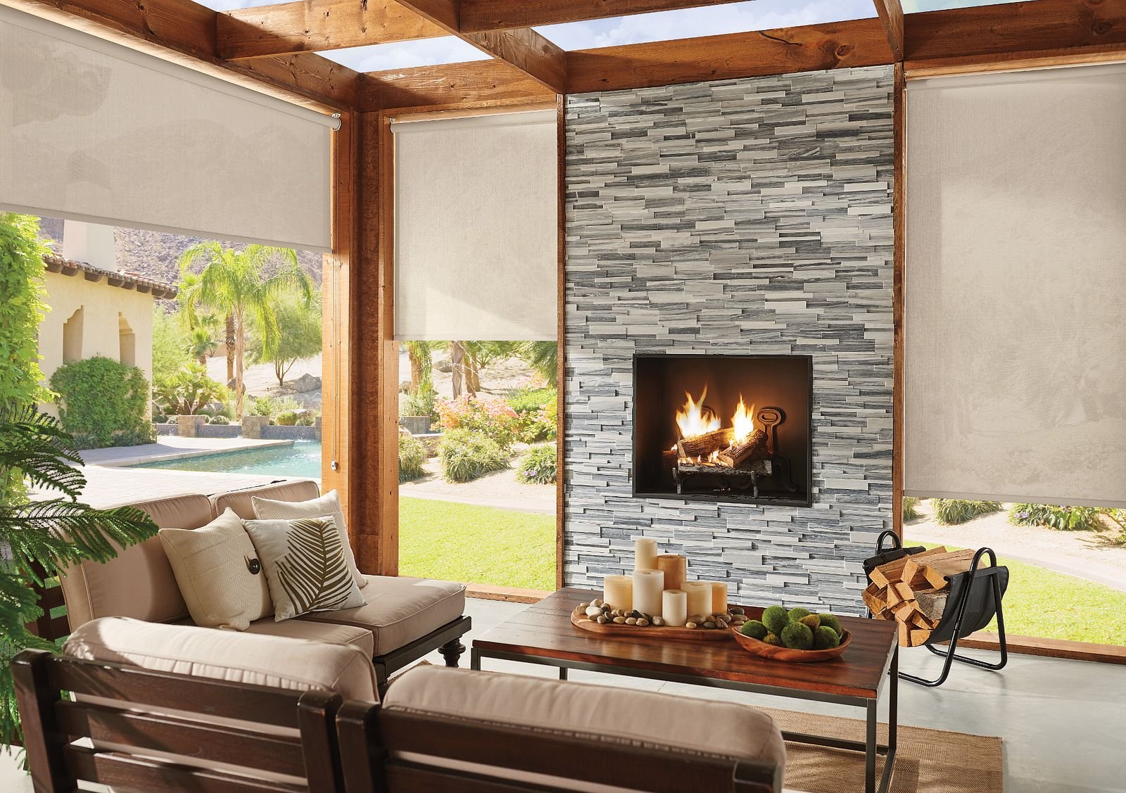 Solar shades on a patio - Pearl & Co Solar shades lowered on a covered patio with a stone fireplace, outdoor sofas, palm trees and a pool