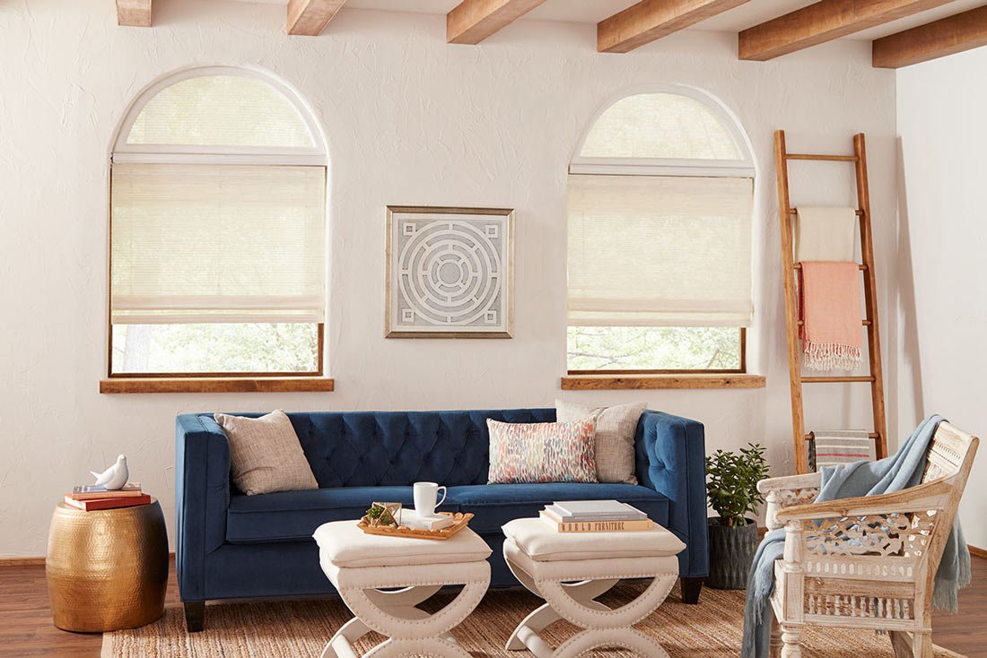 Manh Truc Woven Shades - Pearl & Co Woven wood shades on two arched windows in a white living room with wood ceiling beams and a navy sofa