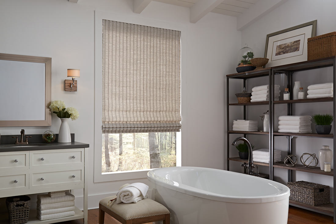 roman-shades - Pearl & Co Tan hobbled Roman shade on a bathroom window behind a freestanding white tub and towel shelves